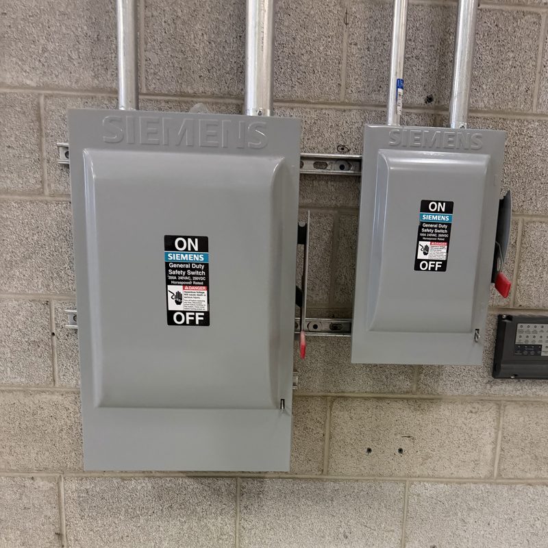 Two grey Siemens safety switches of different sizes on a block wall, fed by conduit from above.