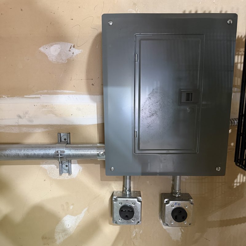 A grey subpanel on a wall with conduit running to two metal boxes holding 240 volt outlets.